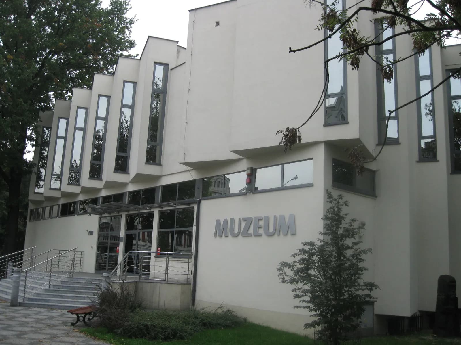 A modern, light-colored building with large vertical windows, a glass entrance, and the word 'MUZEUM' on its facade.