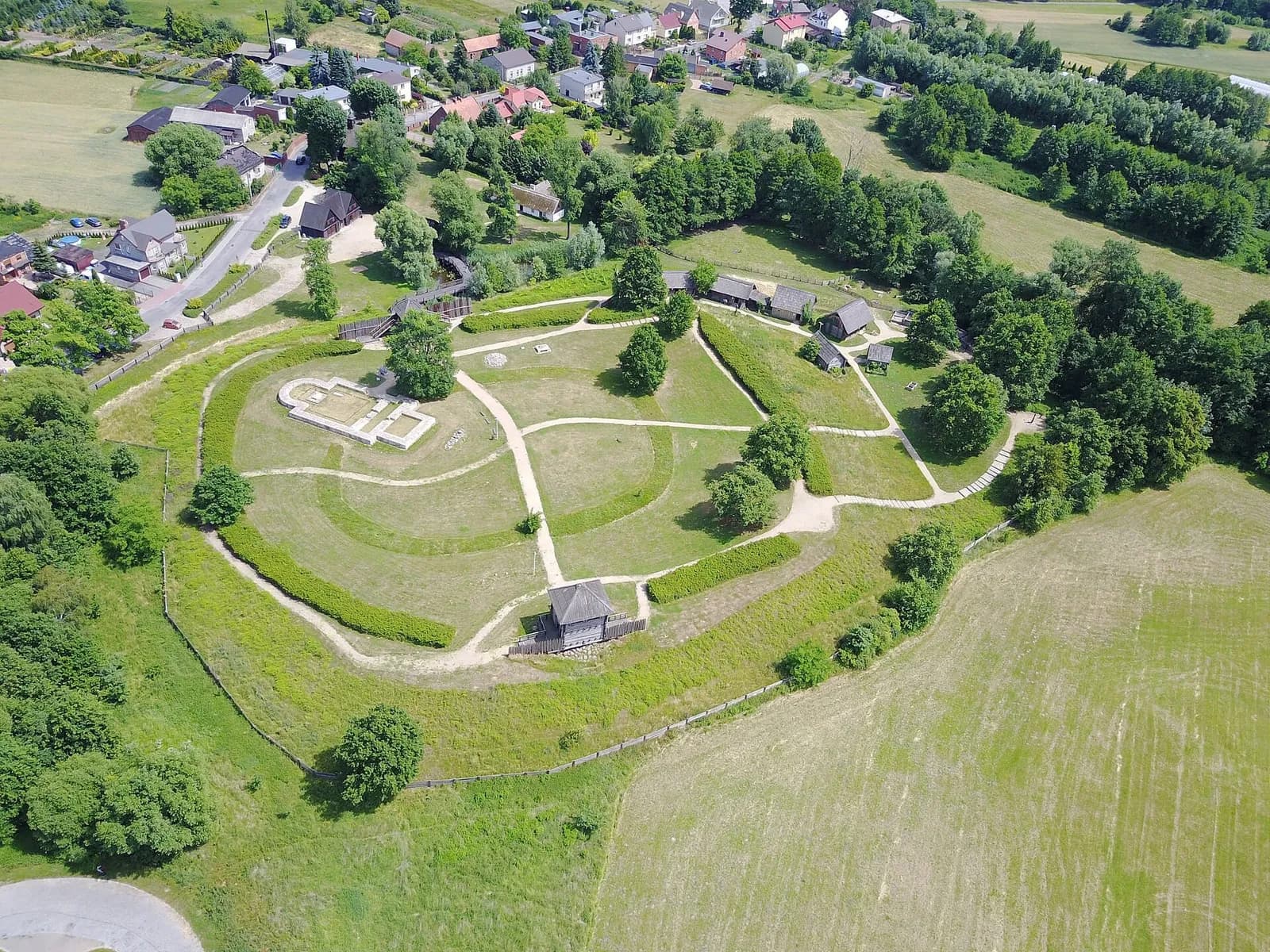 An aerial view captures an ancient settlement on a hill, surrounded by green fields, sparse trees, and a distant village.