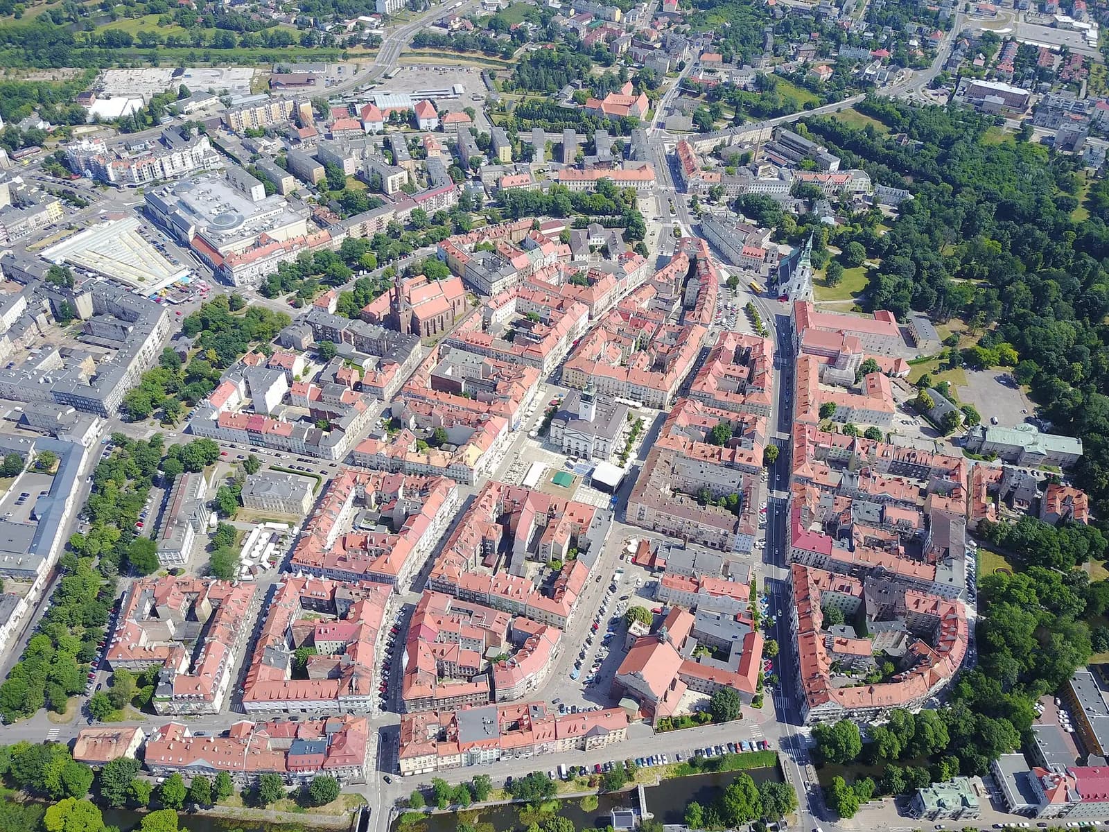 An aerial view of a densely packed city with red-roofed buildings, surrounded by green trees and some larger, more modern structures on the outskirts.